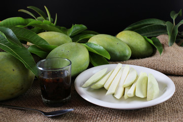 Famous snacks in Thailand, sliced tropical fresh green mango serve with sweet fish sauce, with group of green mangoes and green leaves on burlap sack and black background