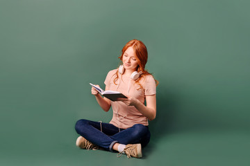 Red-haired young woman with headphones reads a book, sitting on an isolated on green background...