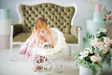 Fototapeta na wymiar Little pretty smiling blonde 3 years old girl in pink dress putting gray and white furry rabbit in white baby carriage. Child and bunny. Spring easter holidays. Friendship between child and animal