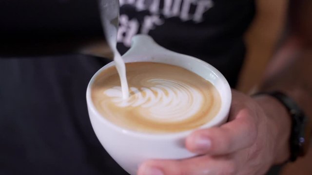 Man pours foamy steamed milk froth into hot cappuccino and creates latte art. Male serves coffee at a rustic cafe shop. Real time stock footage.