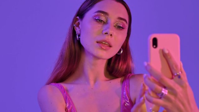A cheerful woman with fashion glitter makeup is making selfie photos isolated over violet neon background