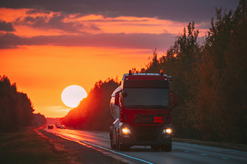 Red Truck Or Tractor Unit, Prime Mover, Traction Unit In Motion On Road, Freeway. Asphalt Motorway Highway Against Background Of Big Sunset Sun. Business Transportation And Trucking Industry.