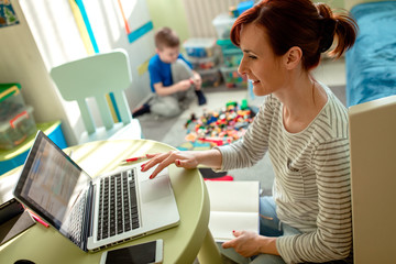 Mother working remotely on laptop while taking care of her son playing with toys in his room.