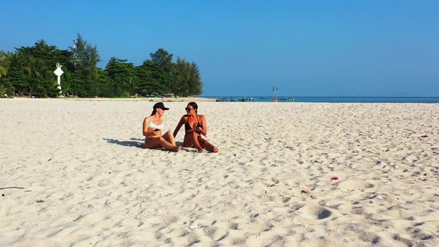 Young women in bikini sunbathe on white sandy beach with no people, smiling and talking with smartphone messages on a sunny day in Cambodia