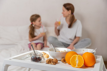 Obraz na płótnie Canvas Happy mom and daughter have healthy breakfast on bed in a light bedroom on a sunny morning. A table with breakfast in the foreground and mom and daughter in defocus. Healthy food concept. Good mood