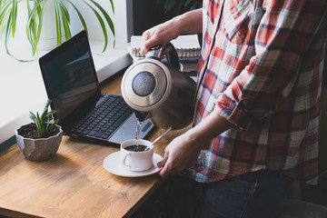 A women pours hot coffee or tea, works on a digital laptop at home, sits at a table. The concept of distance work