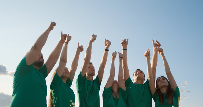 Group of happy young people in green T-shirts raise their hands up and show thumbs up, on the background of blue sky in yellow grass.