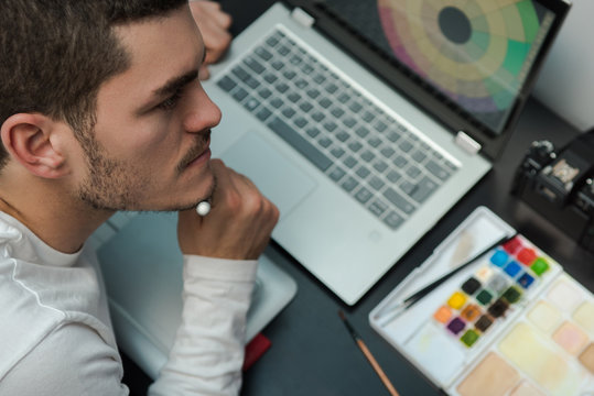 Top view of designer thinking on the workplace hold the electronic work pen. Desktop with laptop, camera, watercolors on a dark table.