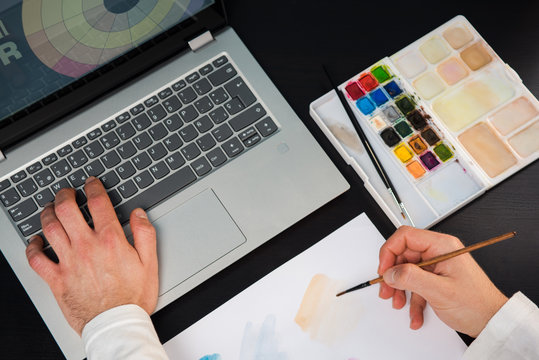 Young man designer painting with watercolors and using the laptop to create. Concept graphic designer studio on a wood table.
