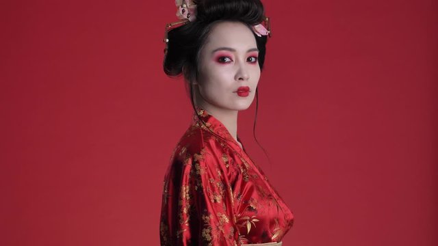 An amazing beautiful young geisha woman in traditional japanese kimono is posing while turning around isolated over red background