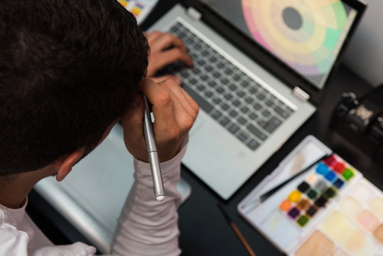 Top view of designer thinking on the workplace hold the electronic work pen. Desktop with laptop, camera, watercolors on a dark table.