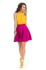 Cute Smiling Walking Woman In Vibrant High Heels, Pink Mini Skirt And YellowTop. Front View.