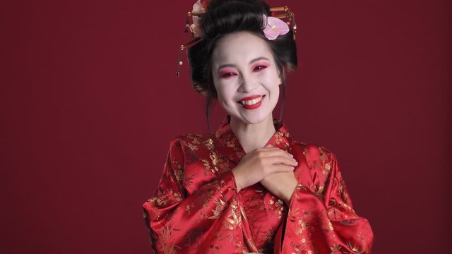 A lovely smiling young geisha woman in traditional japanese kimono is sending an air kiss to the camera isolated over red background