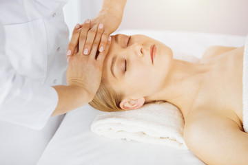 Fototapeta na wymiar Beautiful caucasian woman enjoying facial massage with closed eyes in sunny spa salon. Relaxing treatment in medicine and Beauty concept