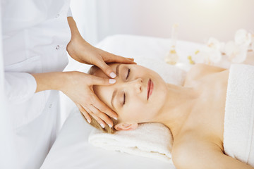 Obraz na płótnie Canvas Beautiful caucasian woman enjoying facial massage with closed eyes in sunny spa salon. Relaxing treatment in medicine and Beauty concept