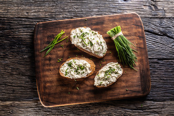 Slices of bread with a cottage cheese spread, freshly cut chives, and a bunch of chive aside placed on a dark brown cutting board and vintage background - obrazy, fototapety, plakaty