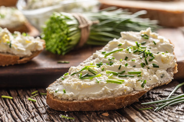 Slices of crusty bread with a cream cheese spread and freshly cut chives on a vintage wooden cutting board