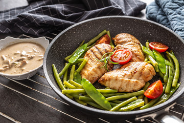 Fried chicken breasts and veg in a frying pan with a creamy mushroom sauce on the side, placed on a black background with a towel and a potholders