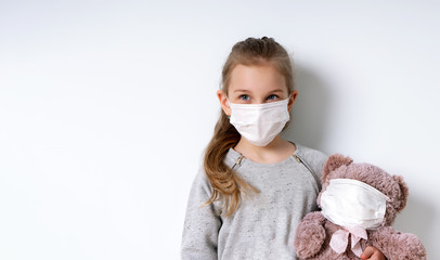 Blonde little model in gray jumper and medical mask, posing isolated on white. She holding toy teddy bear which is also in mask. Coronavirus. Quarantine. Pandemic COVID-19. Close up, copy space
