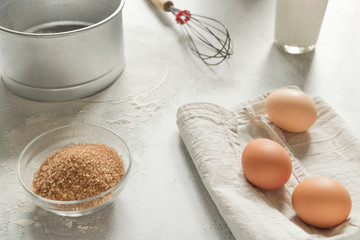 Eggs on a white dish-cloth, whisk, glass of milk, brown sugar and flour sieve