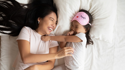 Above top view cheerful Asian mother tickling play with little toddler daughter in pink eye sleeping mask on head, happy family lying in bed awakened after night sleep feels refreshed and vivacious