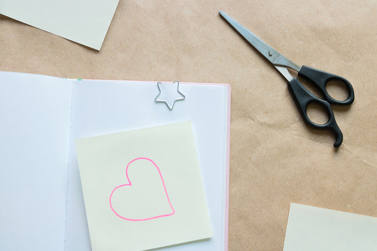 A pair of cissors and a sticky note with a picture of a heart, placed on an opened notebook