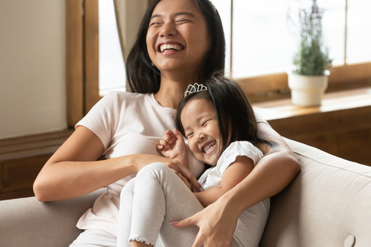 Lively asian young mother tickling little daughter enjoy free time active weekend together, wearing casual comfortable home clothes kid girl tiara accessory, beautiful happy family having fun concept