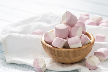 Fototapeta na wymiar Wooden bowl full of pink and white marshmallows with some scattered around on a white table cloth