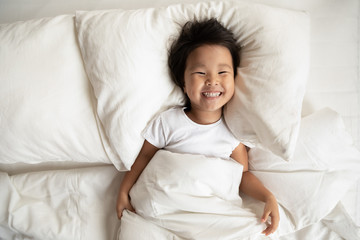 Top view in bed fresh sheets comfortable mattress soft pillow white bedding lies small asian girl has wide charming toothy smile looks at camera, kid woke up, enough healthy sleep, feels active lively