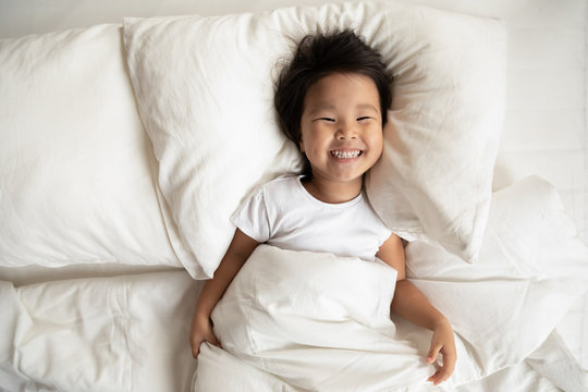 Top View In Bed Fresh Sheets Comfortable Mattress Soft Pillow White Bedding Lies Small Asian Girl Has Wide Charming Toothy Smile Looks At Camera, Kid Woke Up, Enough Healthy Sleep, Feels Active Lively