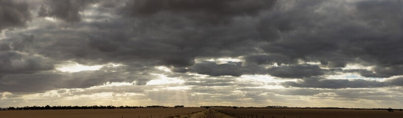 Obraz premium panoramic of a dramatic cloud filled late afternoon sky stretching over large farm fields in rural Victoria just after harvest with a dirt road down the middle.