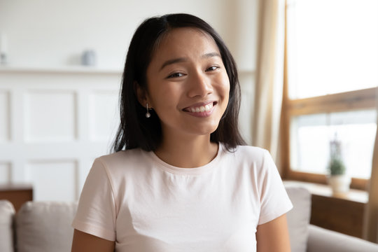 Head shot portrait Vietnamese millennial attractive woman sit on couch in living room alone, smiling looking at camera webcam, make video conference call distant interaction using modern tech concept