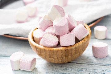 Fototapeta na wymiar Close up of a wooden bowl full of pink and white marshmallows with some scattered around on a white table cloth, dark tray and white wooden table