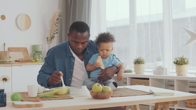 Medium shot of African man sitting at kitchen table, holding curly-haired toddler, feeding him with healthy fruit puree