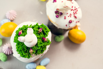 Orthodox Easter cakes decorated with cream in the form of grass and rabbit figurines made of white chocolate on a gray background next to painted eggs. Easter food concept. Idea for kids