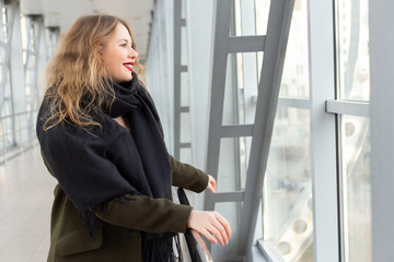 Stylish fashion portrait of blonde woman. Posing in the city. Beautiful girl in autumn green coat and black scarf poses in an overhead pedestrian crossing.