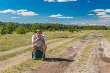 Mature man with old suitcase sitting on an country road and unsuccessfully waiting for any bus to pass by