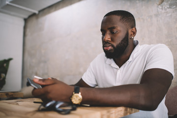 African American bearded student using cellphone application for online web banking and text communication, millennial hipster guy 20s connecting to 4g wireless for networking social website