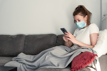Woman wearing medical face mask holding phone while sitting on couch. Covid-19 outbreak pandemic. Stay home concep.