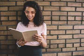 Pretty African American female teenager in stylish pink sunglasses standing near promotional background and reading book, cheerful dark skinned hipster girl smiling near brick wall enjoying leisure