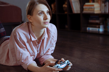 Young attractive woman with dark hair and eyes plays computer game, laying on the floor of her room. Isolation time spending, leisure on quarantine period. Controller in her hands, indoors, copy space