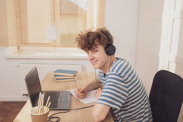 A nice teenage boy in headphones uses the laptop and learns at the desk in his room. He takes notes with a pencil. Distance learning because of the epidemic coronavirus