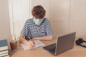 A nice teenage boy in a protective face mask uses the laptop and learns at the desk in his room. He takes notes with a pencil. Distance learning because of the epidemic coronavirus