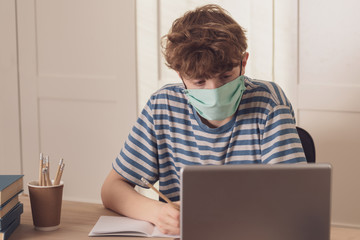 A nice teenage boy in a protective face mask uses the laptop and learns at the desk in his room. He takes notes with a pencil. Distance learning because of the epidemic coronavirus