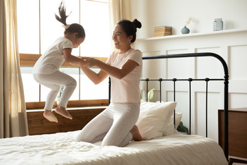 Asian mother holding hands of little toddler daughter while she jumping on bed. Family spend...