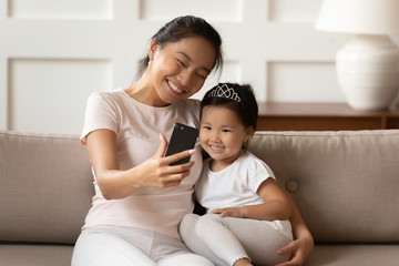 Cheerful Asian mother cuddles little sweet daughter holding smartphone making video call talk to...