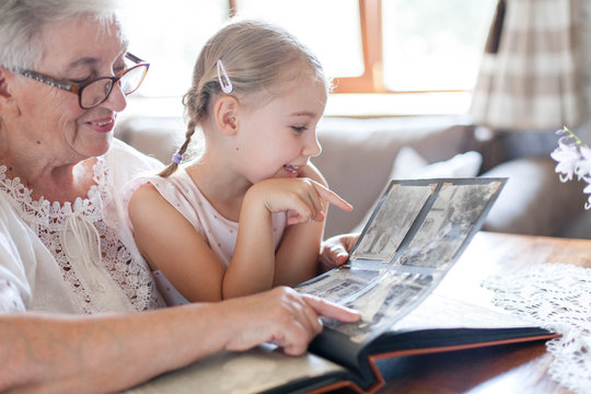 Happy family watching old photo album at home together. Senior woman showing child black and white vintage photos. Retired person memories. Leisure of grandmother and kid. Lifestyle authentic moment.