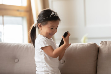 Preschool toddler vietnamese adorable girl sit on couch holding smart phone looking at screen smiling watching cartoon, using educational on-line application for kids, children and modern tech concept