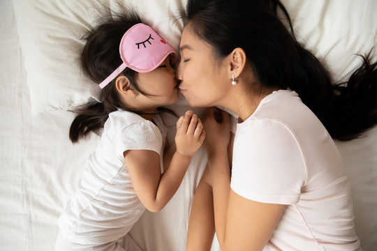 Sweet tender moment of mom kiss child close up top view. Loving mum puts child to healthy day night sleep, asian family lying in bed, kid girl wears pink sleeping eyemask. Love health care concept