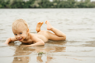 The boy, about two years old, lying naked on the beach on the river beach getting a sun tan alone on a beautiful beach on a clear day.Touching water with hand and looking at camera.